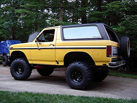 1985 Ford Bronco - This lifted Ford Bronco sits on a driveway, its yellow paint contrasting with the surrounding trees and grass.