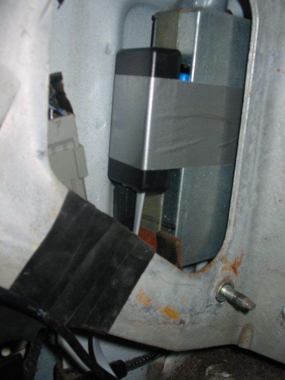 Electronic module secured with silver tape inside a vehicle's compartment. Some wiring and black rubber trim are visible