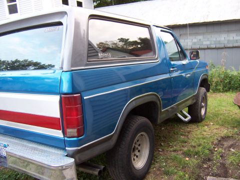 1983 Ford Bronco - Three-quarter rear view of a blue and silver two-tone Ford Bronco parked on grass beside a house, showing the removable fiberglass hardtop,…