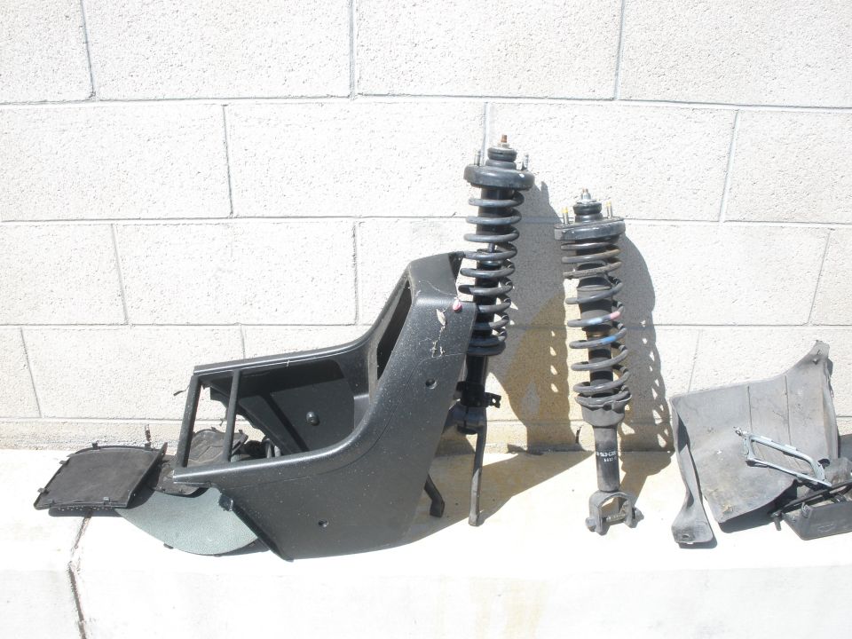 Car parts including a seat frame and two shock absorbers are laid out against a brick wall