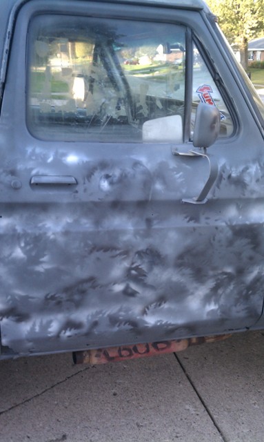 1978 Ford Bronco - The side of a weathered truck door shows primer and rust, with red lettering visible on the exhaust pipe.