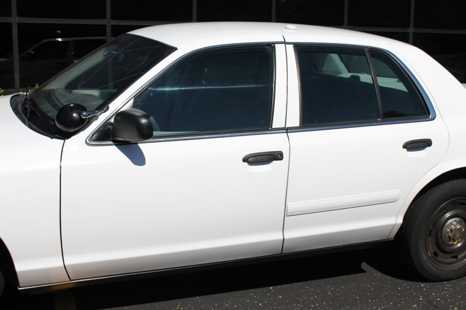White 2003 Ford Crown Victoria sedan, side view, with blacked-out windows and a spotlight attached to the driver's side windshield