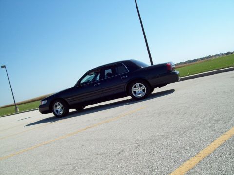 1999 Mercury Grand Marquis - A dark sedan is captured from a 3/4 front angle, parked on a paved surface with a clear blue sky above.