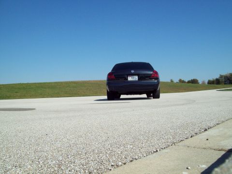 1999 Mercury Grand Marquis - Rear view of a dark sedan on a paved road with a grassy hill in the background under a clear blue sky.