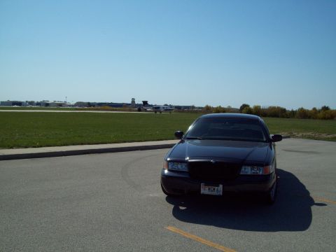 1999 Mercury Grand Marquis - A dark sedan is parked on asphalt with a grassy field and airport in the distance under a clear blue sky.