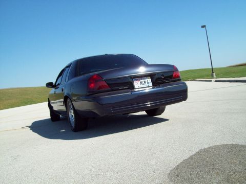 1999 Mercury Grand Marquis - A dark blue sedan is viewed from the rear, angled upwards on a paved surface under a clear blue sky.