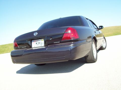 1999 Mercury Grand Marquis - A dark-colored sedan is captured from a low rear quarter angle against a clear blue sky.