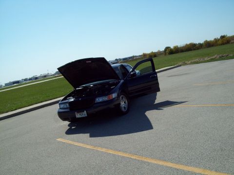1999 Mercury Grand Marquis - A dark sedan sits in a parking lot with its hood and driver's door ajar under a clear blue sky.
