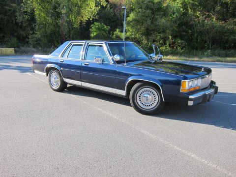 1991 Ford LTD Crown Victoria - A dark blue sedan is parked on an asphalt lot with trees in the background.