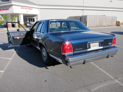 1991 Ford LTD Crown Victoria - A dark blue sedan with its driver's door ajar is parked in a lot outside a building with a drive-thru pharmacy.