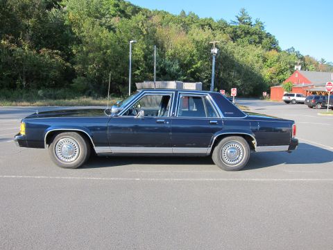 1991 Ford LTD Crown Victoria - A dark blue sedan is parked in an asphalt lot with trees and buildings in the background.