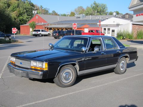 1991 Ford LTD Crown Victoria - A black sedan is parked in a lot with other vehicles and buildings in the background.