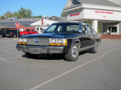 1991 Ford LTD Crown Victoria - This 1980s Ford Crown Victoria is parked outside a drive-thru pharmacy with other vehicles nearby.