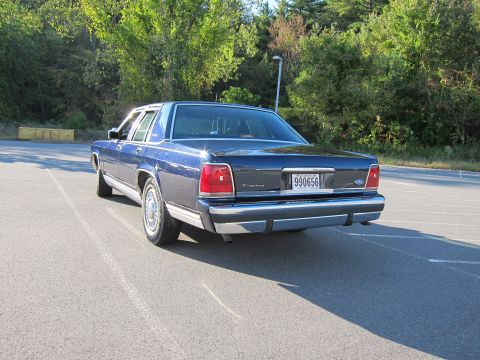 1991 Ford LTD Crown Victoria - Dark blue Ford LTD Crown Victoria sedan photographed from the rear three-quarter in an empty paved lot, showing the full-width taillight pa…