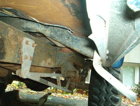 1978 Ford Bronco - Low-angle underbody shot along the driver's side frame rail showing surface rust, bolted body mounts, and a rusted-through rocker with torn…
