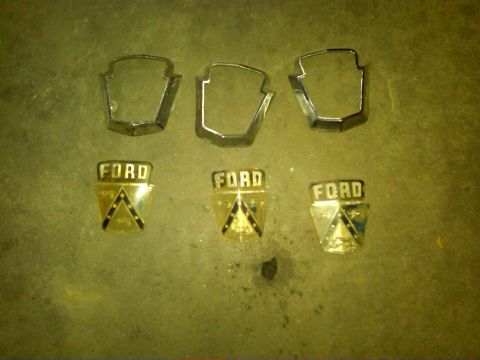 1954 Ford Customline - Three Ford emblems, likely from a 1954 model, are displayed alongside three matching chrome trim pieces.