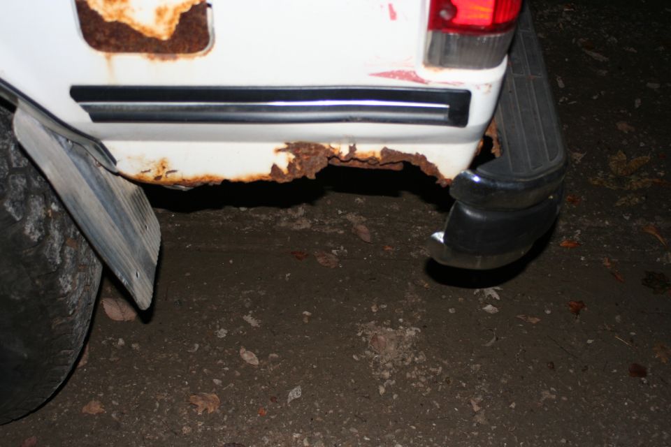 Heavy rust holes on lower rear quarter panel of white Bronco, with black bumper and mud flap, at night
