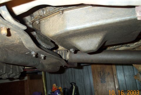 1986 Ford Bronco - From-below view of a truck chassis showing a large stamped-steel transmission or transfer case pan, a rusty driveshaft running along the fr…