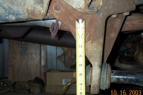 1986 Ford Bronco - A yellow tape measure held vertically against a rusty cast frame crossmember bracket to check roughly six inches of clearance down to the r…