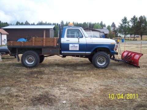 1973 Ford F-250 - Side profile of a blue-and-white 4x4 Ford pickup converted to a wood-sided flatbed dump body, parked on dry grass with pines and a metal sh…