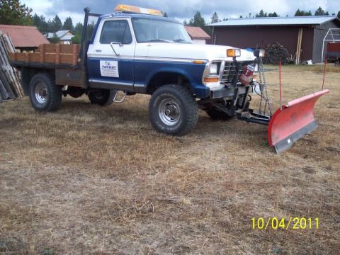 1973 Ford F-250 - Three-quarter front view of a blue-and-white 1970s Ford 4x4 flatbed pickup with wood stake sides, parked on dry grass in front of a dark re…