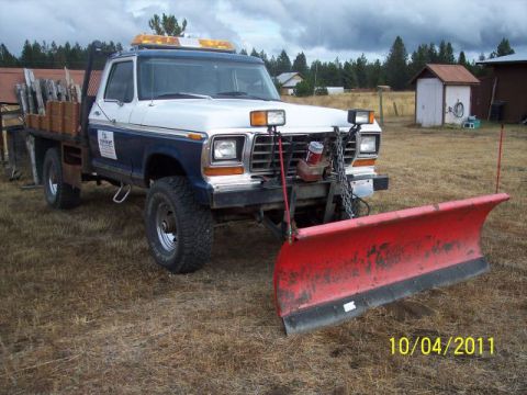 1973 Ford F-250 - Three-quarter front view of a blue-and-white 1970s Ford 4x4 flatbed pickup with wood stake sides and a black headache rack, sitting in a dr…