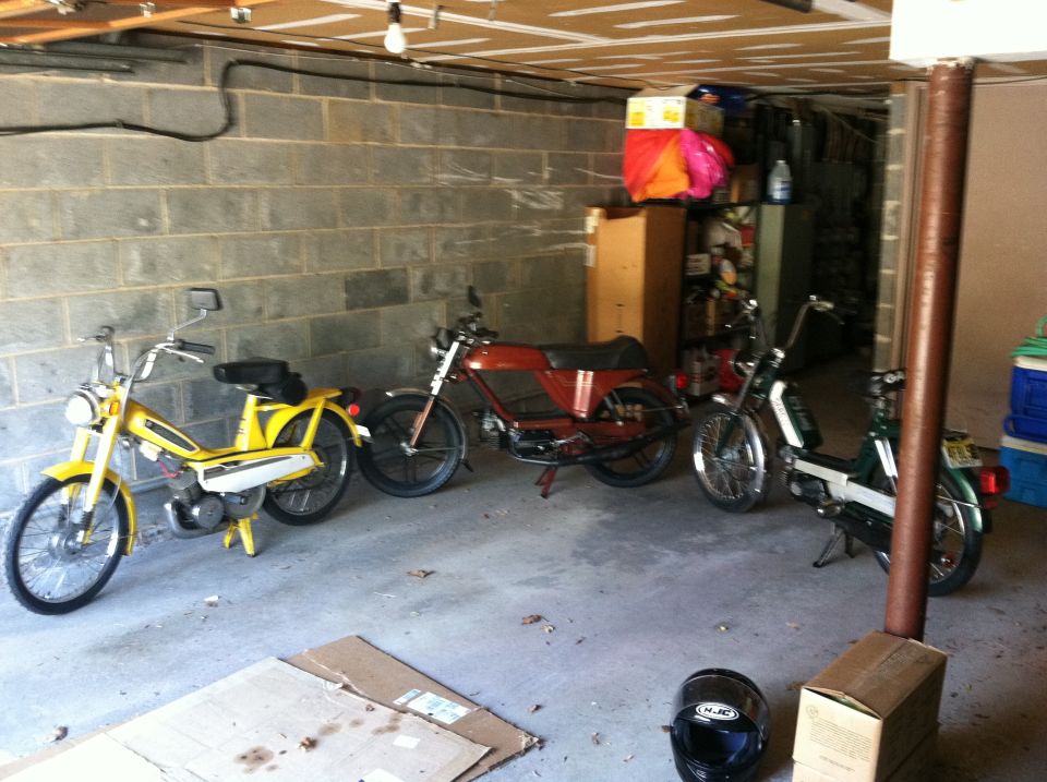 Three vintage mopeds parked in a garage, yellow, red and green