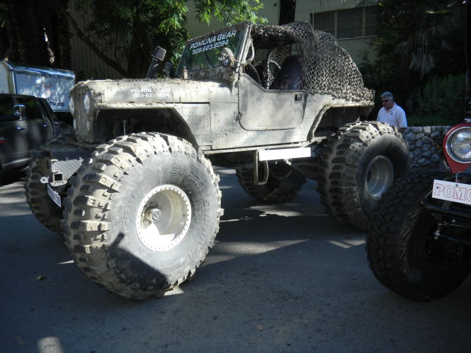 Modified Jeep CJ with large off-road tires and camouflage netting on the roof. Text on door reads 'POMONA GEAR 909.623.8094'. Another vehicle is partially visible to the right