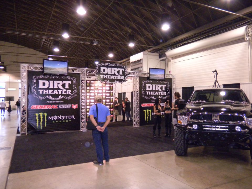 Black off-road truck with large tires and light bar parked next to a 'Dirt Theater' display at an indoor event