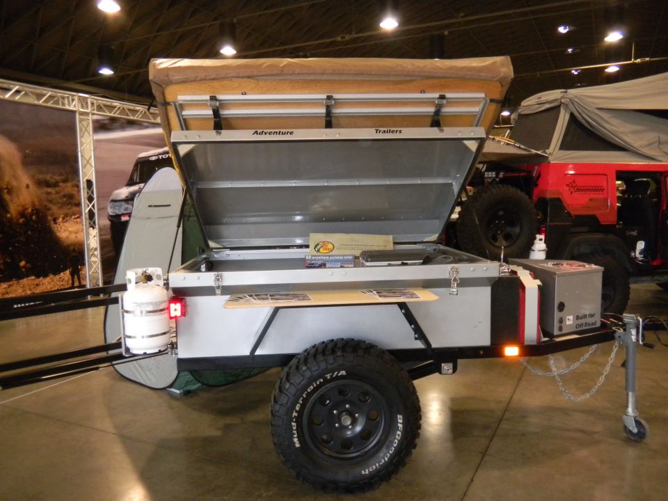 Off-road trailer with open lid, propane tank, and rooftop tent; red Jeep visible behind
