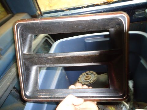 1985 Ford Bronco - A hand holds a dark dashboard trim piece with a wood grain accent, showing a glimpse of a blue interior behind it.
