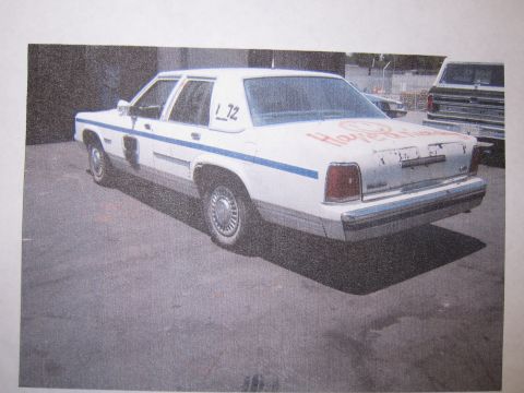 1991 Ford LTD Crown Victoria - printed photograph shows a white 1980s sedan with a blue side stripe and orange spray-painted graffiti, viewed from the rear.