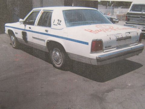 1991 Ford LTD Crown Victoria - printed photo of a white 1980s sedan with blue stripe and orange spray-paint, parked in an indoor lot.