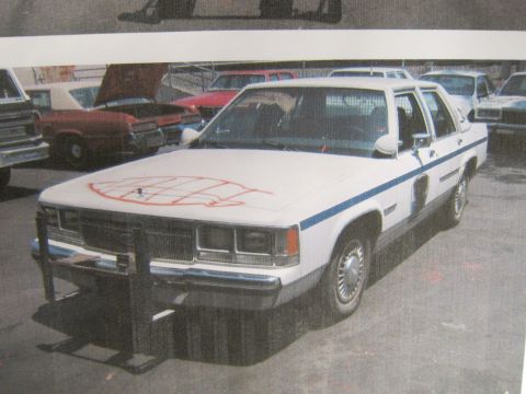 1991 Ford LTD Crown Victoria - printed photo shows a white Ford sedan with a front push bar and orange spray-paint scrawl across the hood.
