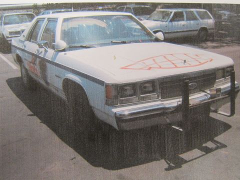 1991 Ford LTD Crown Victoria - printed photograph of a white Ford sedan with a push bar and orange spray-paint on the hood parked among other cars.