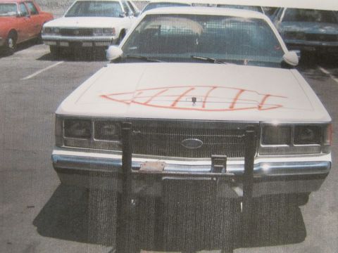 1991 Ford LTD Crown Victoria - printed photo of a white sedan with a front push bar and orange spray-painted markings across its hood in a parking lot.