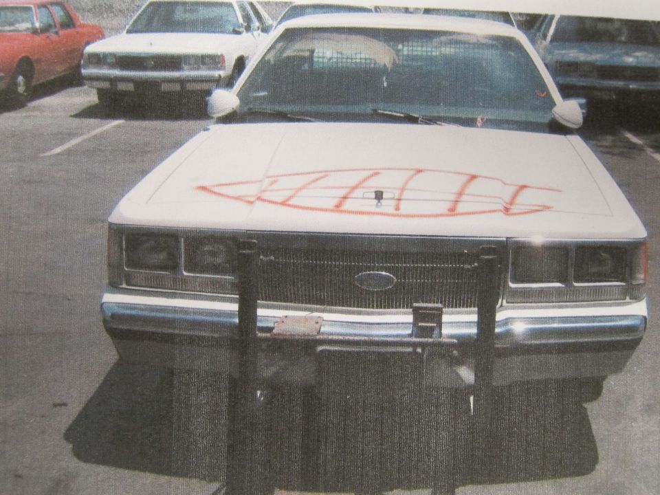 Printed photo of white sedan front with push bar and orange spray-paint on hood