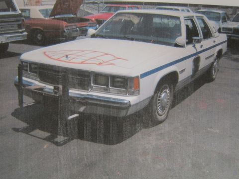 1991 Ford LTD Crown Victoria - printed photograph of a white Ford sedan with a front push bar and orange spray-paint on the hood among parked cars.