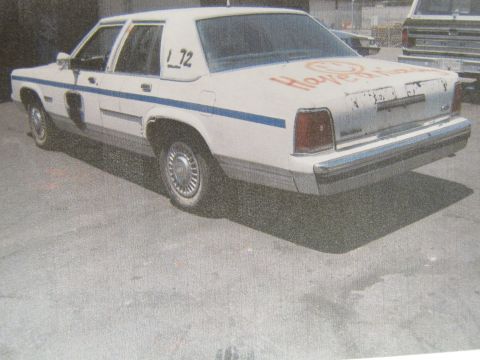 1991 Ford LTD Crown Victoria - printed photo shows a white sedan with a blue stripe and orange spray-paint scrawl, viewed from the rear left in a lot.