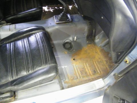 1970 Ford Mustang - The interior floor of a vehicle shows significant rust and wear, with black vinyl seats and a manual transmission shifter visible.