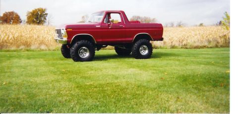 1978 Ford Bronco - A red 1970s Ford Bronco with a lifted suspension, large off-road tires, and chrome wheels, shown in side profile parked on a grassy field i…