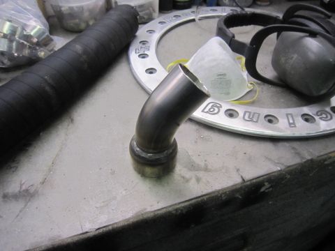 1974 Ford Bronco - A metal exhaust pipe elbow sits on a workbench beside a large ring that reads 'Go Fast!' and other automotive parts.