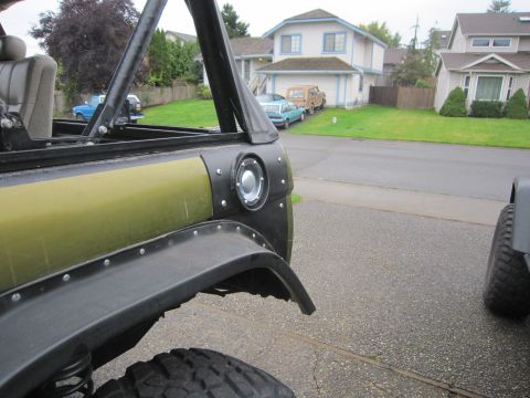 1974 Ford Bronco - Close-up view of the rear quarter panel and taillight assembly on an olive green SUV, with other vehicles visible in the background.