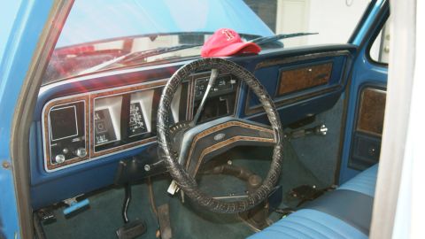1978 Ford Bronco - The dashboard of an older blue truck, wood-grain trim and a worn steering wheel, with a red cap on the dash.