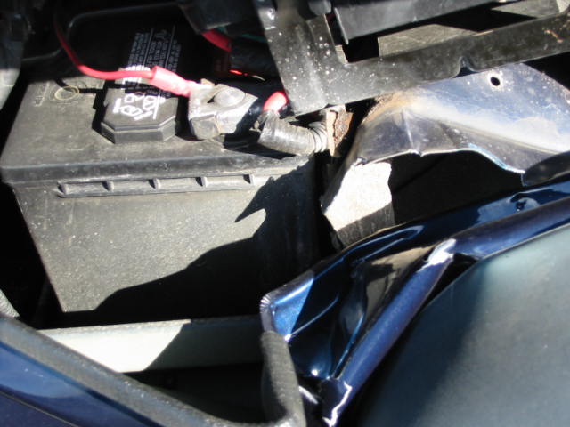 Close-up of torn, crumpled dark-blue sheet metal beside the battery after a wreck