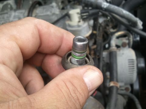 1994 Ford Bronco - A hand holds a metal AC system part with green O-rings against a blurred engine background.