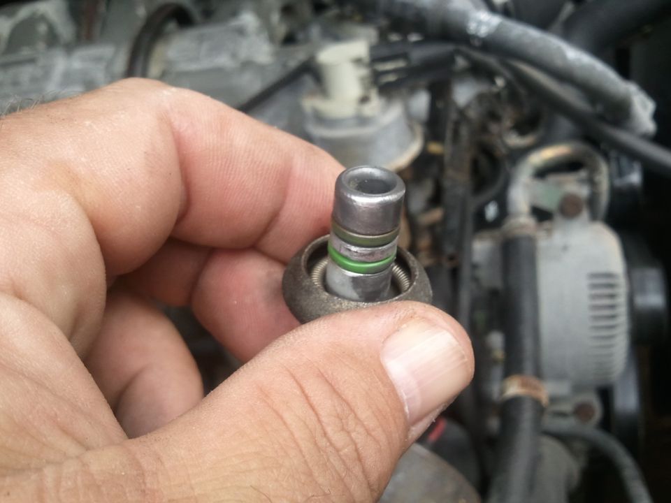 Hand holding a metal AC system part with green O-rings, engine components in background
