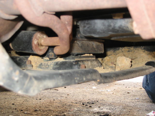 Underside of vehicle showing rusty shackle bolt and frame components over dirt ground
