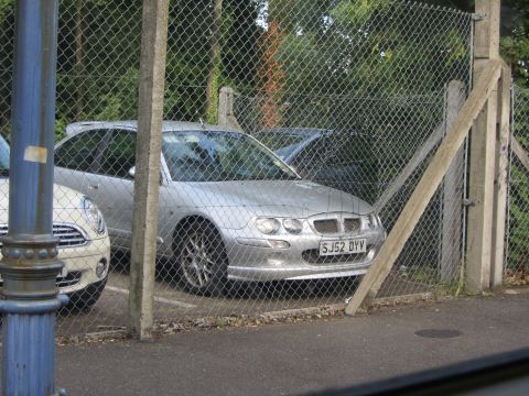 1968 Ford Mustang - A silver MG ZS hatchback is parked behind a chain-link fence, with a white Mini Cooper visible to its left.
