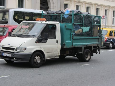 1968 Ford Mustang - A white Ford Transit flatbed truck is seen on a city street, carrying a load of trash bags in its cage.